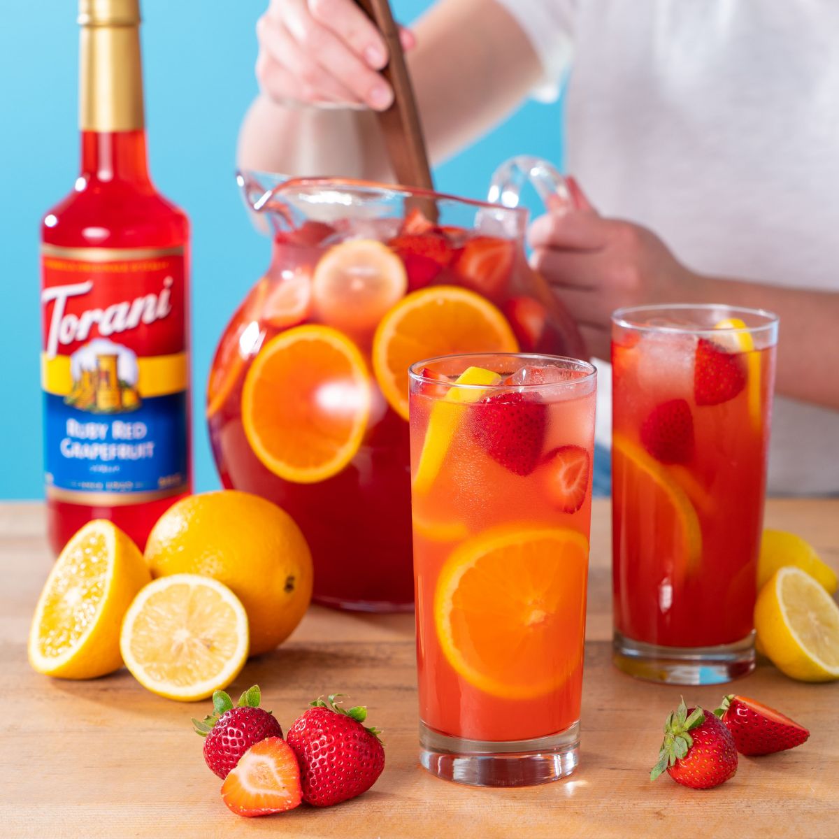 bottle of torani ruby red grapefruit syrup with two sans-gria glasses and a pitcher of sans-gria being made