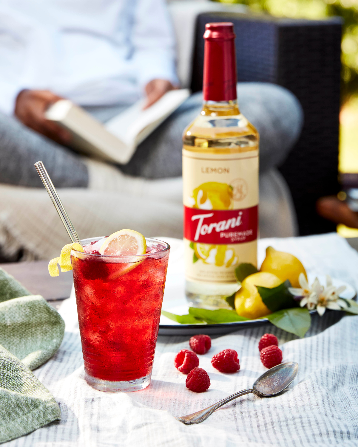 A table with a glass of Lemon Raspberry Iced Tea with a bottle of Torani Puremade Lemon and fresh lemons and raspberries. A woman sits in the background reading a book on their back porch on a sunny afternoon.